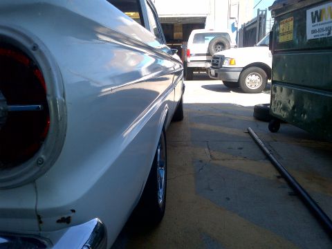 1965 Ford Mustang - A white classic car is parked next to two trucks and a dumpster in an outdoor lot.
