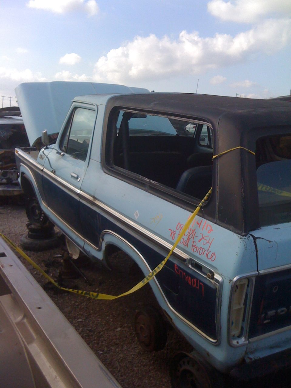 light blue SUV with black top, hood up, parked in a junkyard. Yellow tape on rear quarter panel