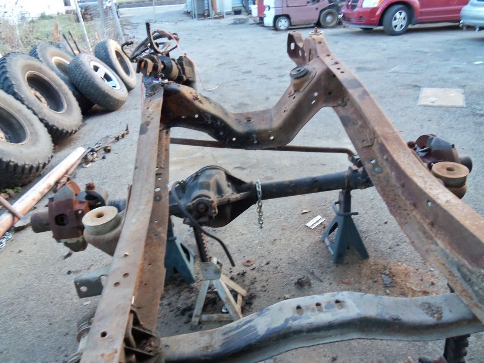 Rusty vehicle frame and rear axle assembly on jack stands, with tires stacked nearby