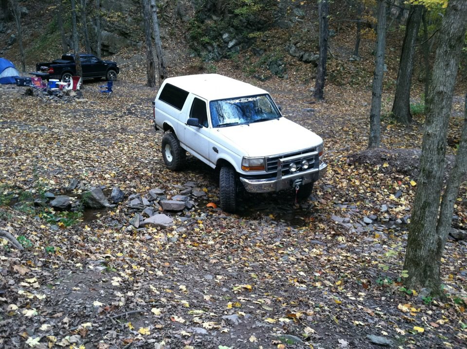 White SUV with lifted suspension parked in a forest campsite. Another black truck is visible in the background