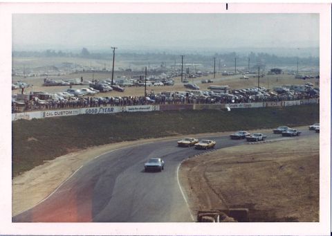 1973 Ford Mustang - Race cars compete on a track with spectators and a large parking area in the background.