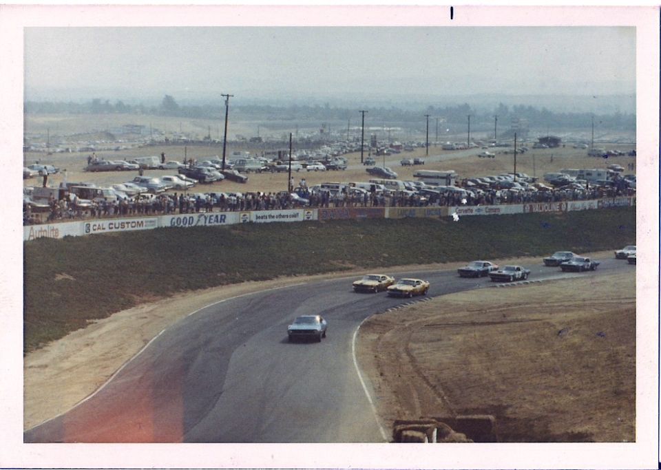 Race cars on a track with spectators and parked cars in the background. Visible text includes 'GOOD YEAR'