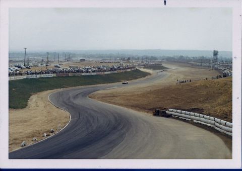1973 Ford Mustang - A race track scene with numerous parked cars and spectators lining the course, with a single car visible on the track.