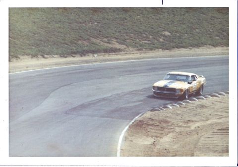 1973 Ford Mustang - A yellow race car with black stripes navigates a turn on a paved track.