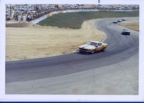 1973 Ford Mustang - A yellow race car navigates a curve on a race track, with spectators and other vehicles in the background.