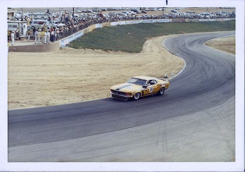 1973 Ford Mustang - A yellow race car with the number 16 on its door navigates a curve on a race track, with spectators and parked cars visible in the backgrou…