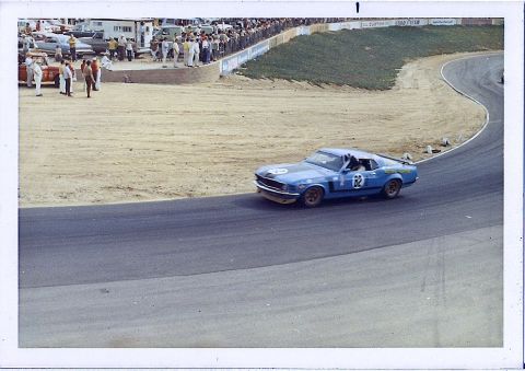 1973 Ford Mustang - A blue race car with racing decals navigates a turn on a sunny day at the track.