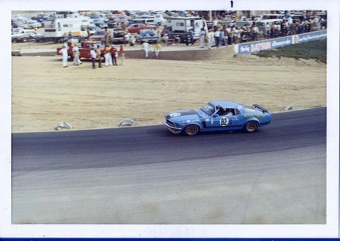 1973 Ford Mustang - A blue race car with number 82 speeds around a track, with spectators watching from the stands.