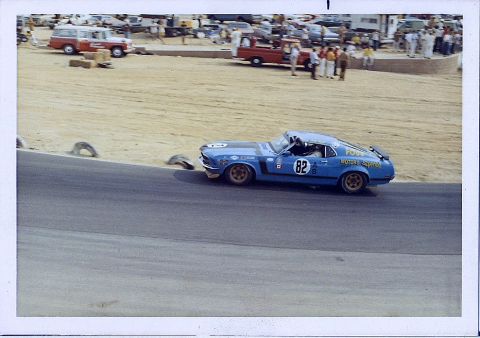 1973 Ford Mustang - A blue race car with number 82 navigates a turn on a track, with spectators and other vehicles in the background.