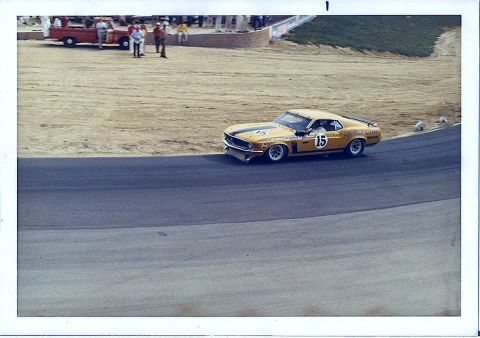 1973 Ford Mustang - A yellow 1970 Boss 302 Mustang race car, number 15, navigates a turn on a track, with a red pickup truck and spectators in the background.