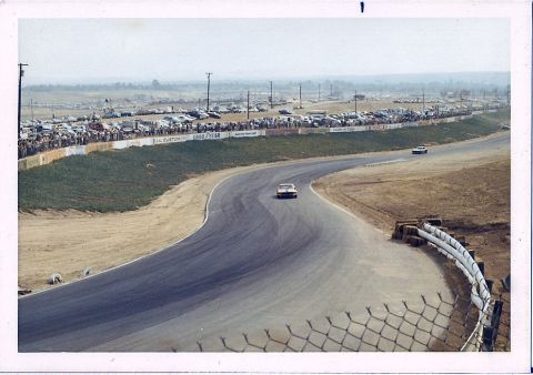 1973 Ford Mustang - Cars compete on a race track, with a crowd watching from the sidelines and a lot full of vehicles in the distance.