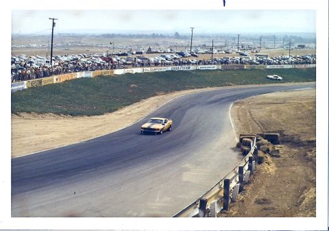 1973 Ford Mustang - A yellow 1970 Ford Mustang speeds around a race track, with a crowd and many parked cars visible in the distance.