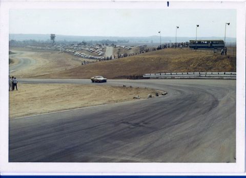 1973 Ford Mustang - A gold 1973 Ford Mustang coupe navigates a turn on a race track, with spectators and parked cars visible in the background.
