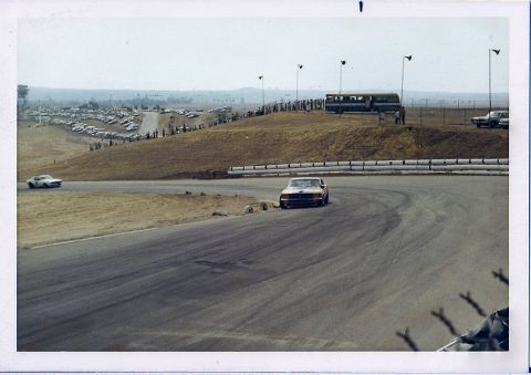 1973 Ford Mustang - A 1973 Ford Mustang navigates a turn on a race track, with a crowd and parked cars visible on the hillside.