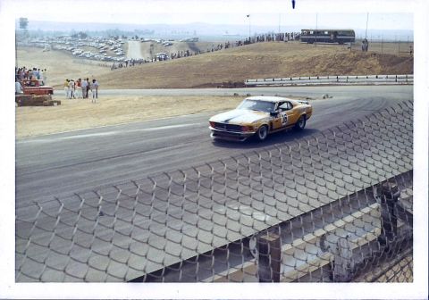 1973 Ford Mustang - A vintage race car speeds around a corner at a track event, with a crowd and parked vehicles visible in the distance.