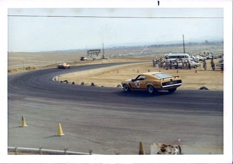 1973 Ford Mustang - A yellow 1970 race car, identified by its body style and number 16, navigates a turn on a sunny day at the track.