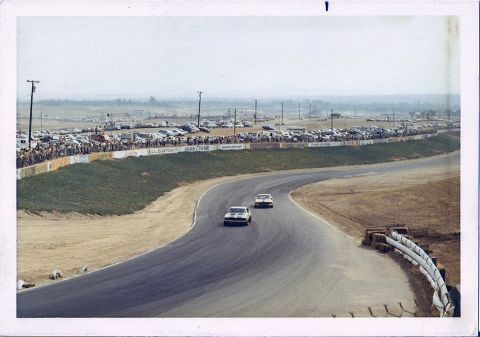 1973 Ford Mustang - Two 1973 Ford Mustangs race on a track with spectators and a large parking lot in the background.