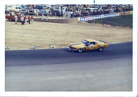 1973 Ford Mustang - A yellow race car speeds around a track, with spectators and other vehicles in the background.