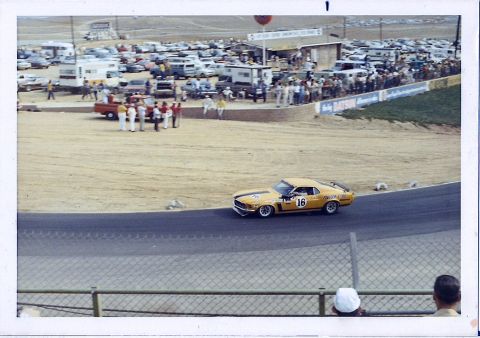 1973 Ford Mustang - A yellow 1970 Ford Mustang Boss 302 race car navigates a turn at a crowded racetrack, with spectators and other vehicles in the background.