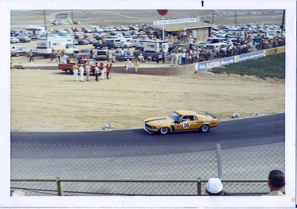 Yellow 1970 Ford Mustang Boss 302 race car with black stripes and number 16 on the door, seen from the front-right at a racetrack