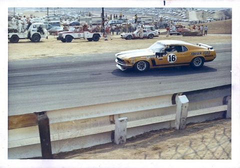 1973 Ford Mustang - A race car speeds around a track, with support vehicles and spectators visible in the background.