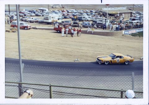 1973 Ford Mustang - A race car speeds around a track, with spectators and parked vehicles visible in the background.