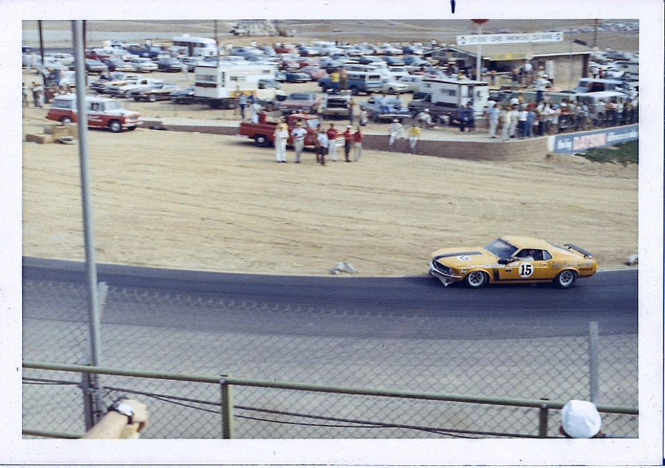 yellow race car with number 15 on track, crowd and parked cars in background