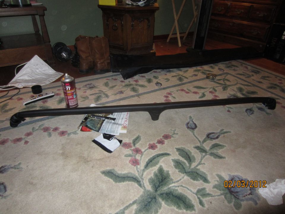 Black car part, possibly a roof rack or trim piece, lying on a floral rug next to a can of spray paint and a remote