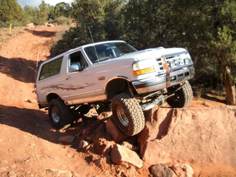 1996 Ford Bronco - A white Ford Bronco tackles a challenging off-road trail with large rocks and steep inclines.