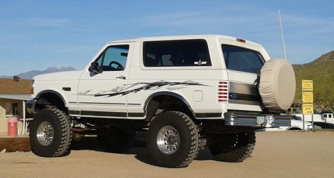 1996 Ford Bronco - A lifted white Ford Bronco with custom graphics sits parked in a dusty lot with mountains in the background.