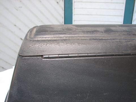 1973 Ford Ranchero - A close-up view of a textured black plastic lid with a metal hinge, set against a white clapboard background.
