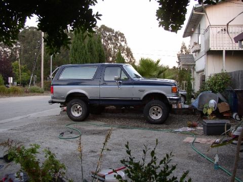1989 Ford Bronco - A two-tone blue and silver Ford Bronco is parked in a residential driveway with a house in the background.