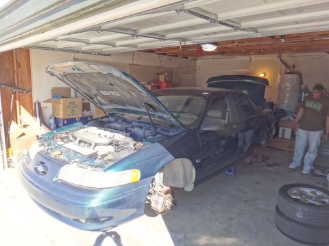 1993 Ford Taurus - A dark green 1993 Ford Taurus coupe is parked in a garage with its hood open and front wheels removed for maintenance.