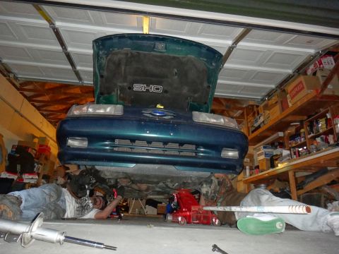 1993 Ford Taurus - Mechanic works on a teal 1993 Ford Taurus SHO in a garage, with the hood up and the car elevated.