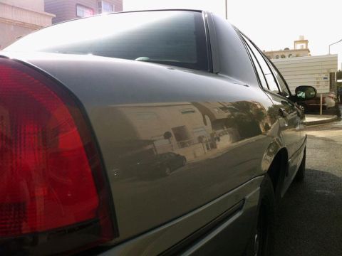 2001 Mercury Grand Marquis - The rear quarter panel of a 2001 Mercury Grand Marquis is visible, reflecting the surrounding buildings and sky.