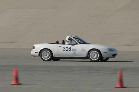 1991 Mazda Miata - A white convertible sports car with racing decals drives on a track, with orange cones visible.