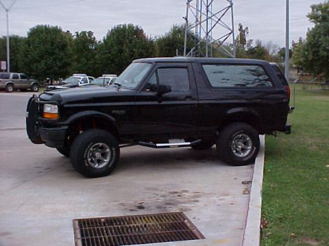 1995 Ford Bronco - A black Ford Bronco is parked on a concrete pad next to a grassy area, with a large metal tower in the background.
