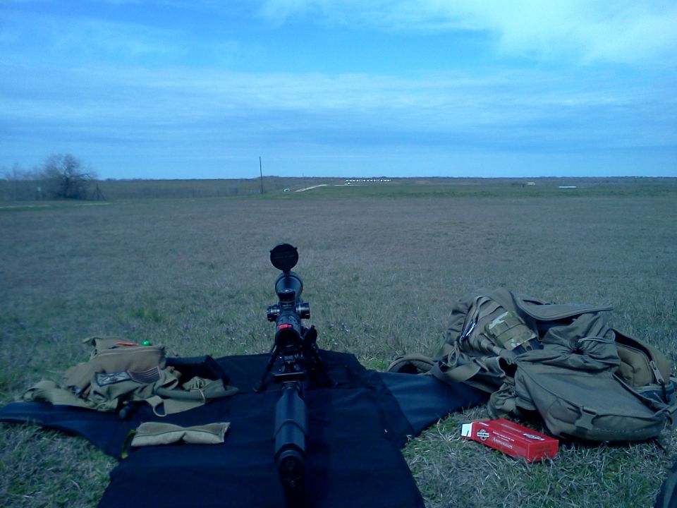 Rifle scope and bipod on a mat in a grassy field with bags and a box of ammunition