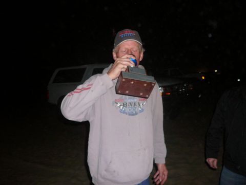 1988 Ford Bronco - A man holds a trophy to his mouth while standing in front of vehicles at night.