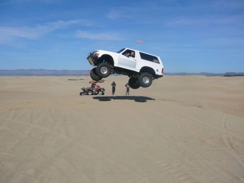 1988 Ford Bronco - A white 1988 Ford Bronco soars through the air during a jump over sand dunes, with an ATV and onlookers below.