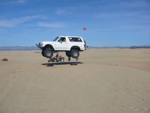 1988 Ford Bronco - White Ford Bronco performs a jump over an ATV in a desert dune area.