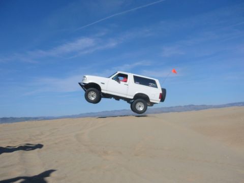 1988 Ford Bronco - A white Ford Bronco is captured mid-air while driving over sand dunes under a clear blue sky.