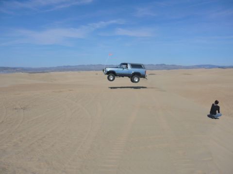 1988 Ford Bronco - A light blue SUV is captured mid-air during a jump over sand dunes, with a lone observer watching from the side.