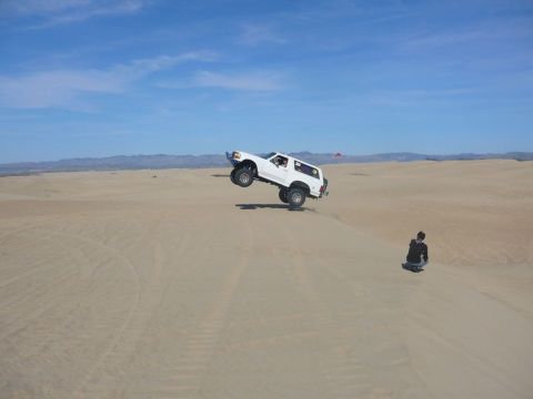 1988 Ford Bronco - A white 1988 Ford Bronco catches air while driving over sand dunes with a person watching in the background.