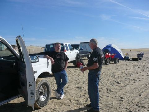 1988 Ford Bronco - Men gathered on a sandy area with vehicles and camping gear under a clear blue sky.