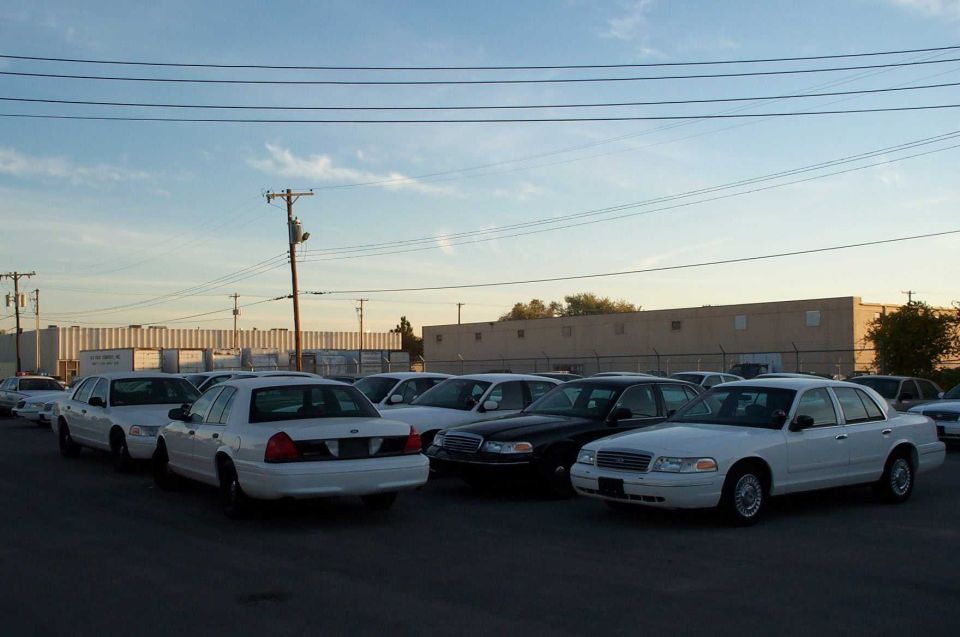 Multiple white and black Crown Victoria sedans parked in a lot at dusk near an industrial building