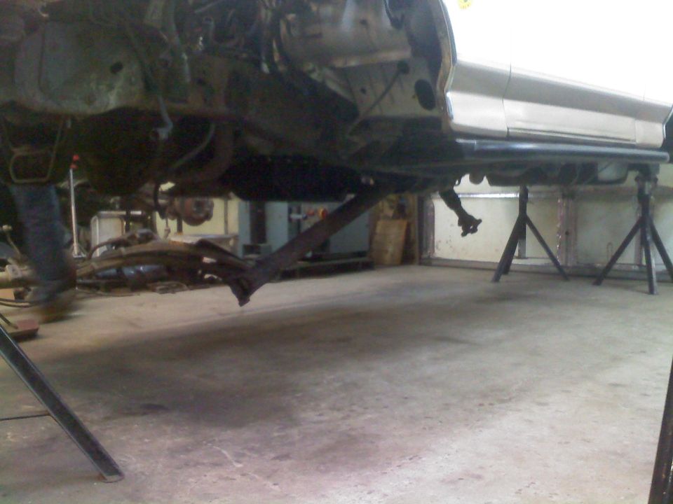 Undercarriage of a vehicle on jack stands, showing suspension components and frame. The vehicle is silver and appears to be undergoing modifications