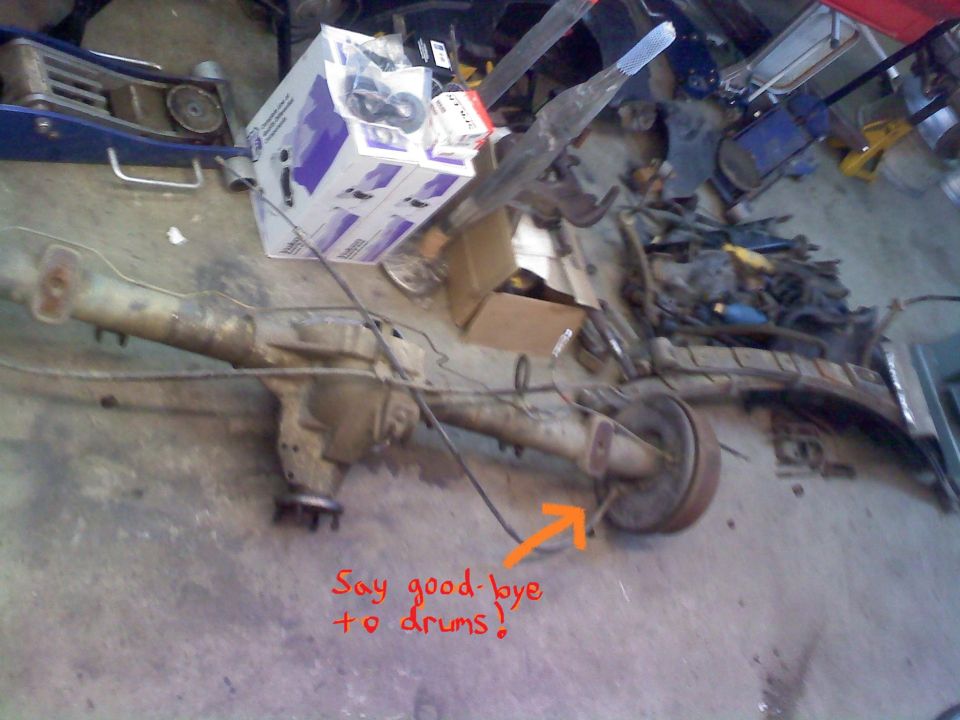 Rear axle assembly with drum brakes and brake lines, on a concrete floor. Text overlay says 'Say good-bye to drums!'
