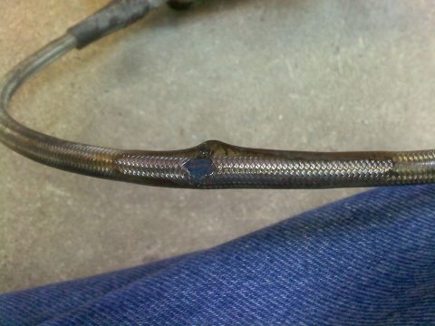 1996 Ford Bronco - A close-up view of a damaged braided hose, showing a hole with a blueish interior, against a backdrop of denim and concrete.