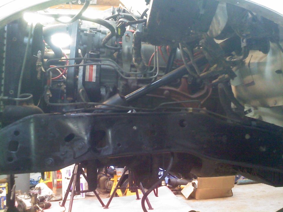 Vehicle engine bay with engine and frame visible, supported by jack stands. Some red and white stripes on a component